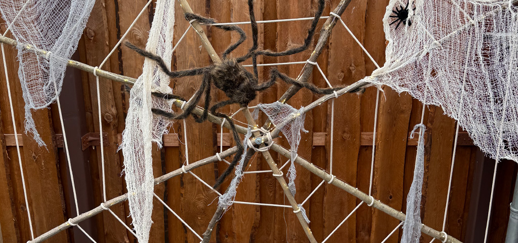 Großes Spinnennetz aus Seilen mit weißen Tüchern und zwei künstlichen Spinnen, auf Holzrahmen im Garten montiert.